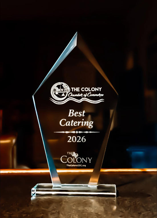 A clear acrylic award trophy with engraved text reading 'The Colony Chamber of Commerce Best Catering 2026' and 'The Colony' branding, displayed on a dark reflective surface against a dim background.