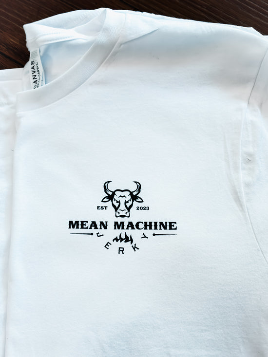 A white short-sleeve t-shirt laid flat on a dark wood surface, featuring a black graphic print on the chest with a bull head illustration and the text 'MEAN MACHINE EST 2023' and 'ERK'.